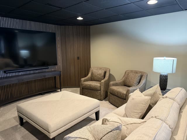 Finished basement media room with a wood slat accent wall, large wall-mounted TV over a console, black ceiling, two leather swivel chairs, an upholstered ottoman and a cream sofa.