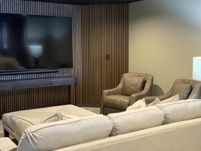 Wood slat accent wall with a wall-mounted TV and soundbar above a dark console table, two leather armchairs and the back of a cream sofa in a finished basement media room.