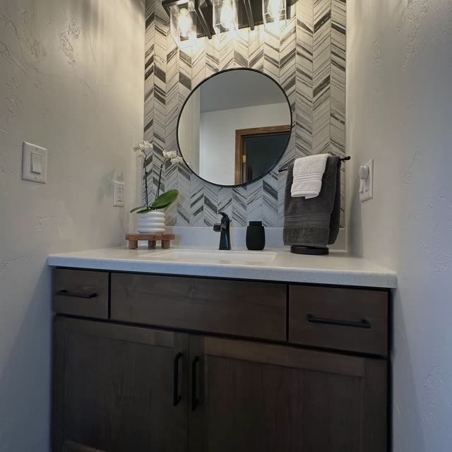 Bathroom vanity in dark stained wood with a white counter, black faucet, round black-framed mirror, a white orchid and a gray and white chevron tile accent wall.