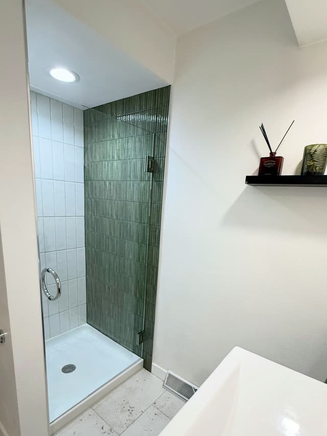 Shower stall with a dark green fluted tile accent wall, white vertical wall tile, a frameless glass door with a chrome ring pull and a white shower base, beside a black floating shelf.