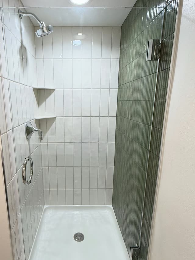 Shower stall with white vertical stacked tile, a dark green fluted tile side wall, chrome shower head and handle, two corner shelves, a frameless glass door and a white shower base.