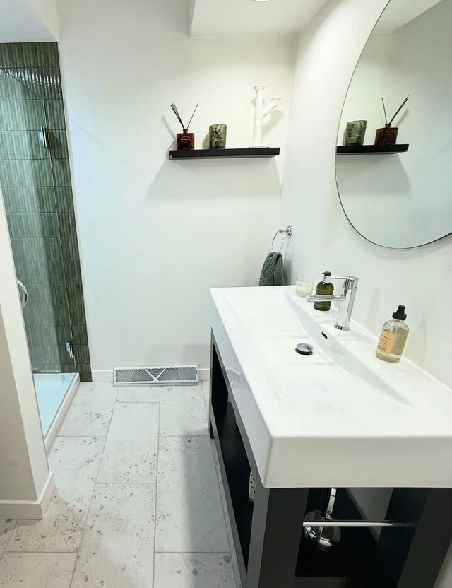 Bathroom with a white rectangular sink on a dark open-leg vanity, a round mirror, a black floating shelf, light speckled floor tile and a green-tiled glass shower in the corner.
