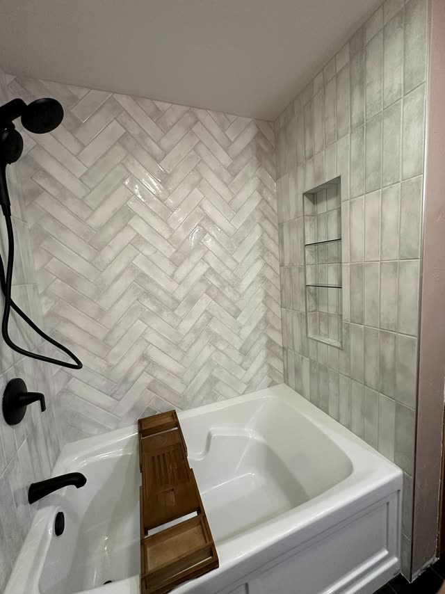 White bathtub with a wooden bath tray, surrounded by gray-white herringbone tile on the back wall, vertical stacked tile on the side wall, a recessed niche with glass shelves and matte black shower fixtures.