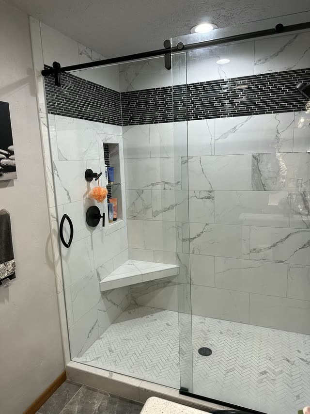 Walk-in shower with white marble-look wall tile, a black glass mosaic accent band, corner bench, recessed niche, herringbone mosaic floor and a frameless sliding glass door on a matte black rail.