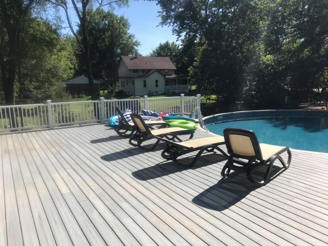 Gray composite pool deck with four lounge chairs facing an above-ground pool, white vinyl railing and tall trees under a blue sky.