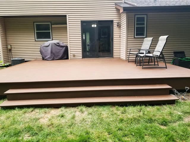 Low brown composite deck with two wide full-width steps behind a tan-sided house, with a covered grill and two patio chairs.