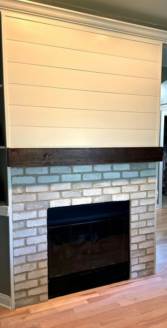 Refaced fireplace with white shiplap above a dark wood beam mantel, a whitewashed brick surround, black glass doors and light hardwood flooring.