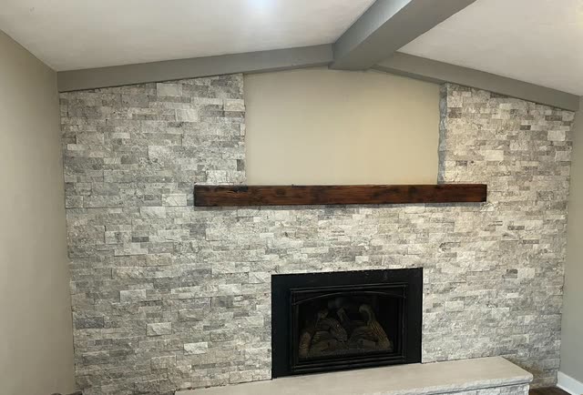 Stacked stone fireplace wall with a black gas insert, a dark rustic wood beam mantel, a raised stone hearth and gray painted ceiling beams.