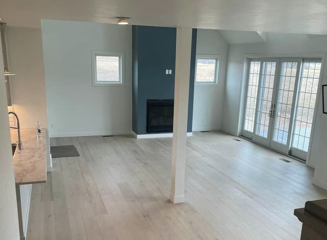 Empty great room with wide-plank light wood flooring, a dark teal fireplace wall, white French doors with grids, and a stair railing in the foreground.