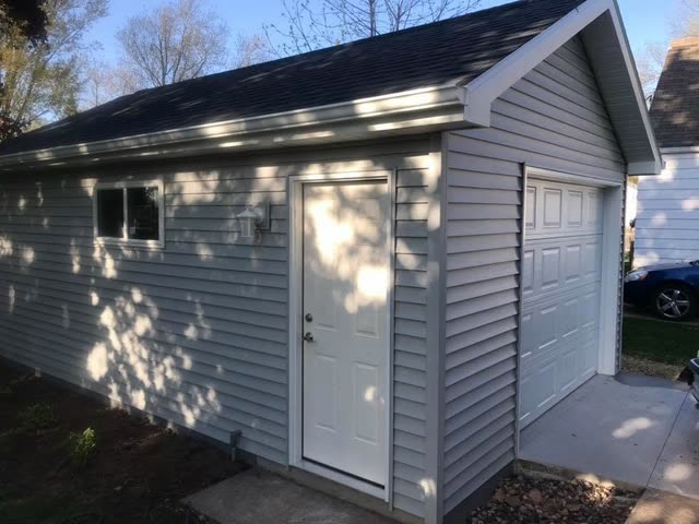 Detached single-car garage with gray vinyl siding, a white service door, a white overhead door and a dark shingle roof.