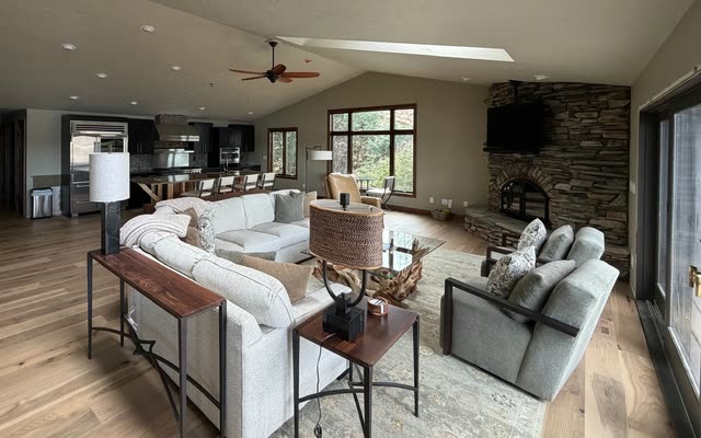 Open great room with wide wood plank flooring, a vaulted ceiling with skylights, a stacked-stone corner fireplace, a white sectional sofa and a dark-cabinet kitchen in the background.