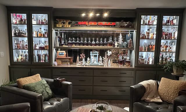 Dark gray custom built-in wall unit with lit glass-door cabinets, open shelves backed with mirrored tile, a bank of drawers below, and leather club chairs around a round wood table in front.