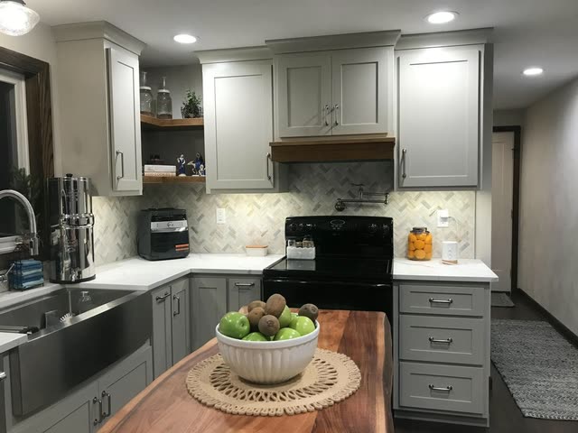 Kitchen with light gray shaker cabinets, white veined countertops, a herringbone marble tile backsplash with a pot filler, a wood-trimmed hood over a black range, a stainless apron-front sink and a live-edge wood island top holding a bowl of fruit.