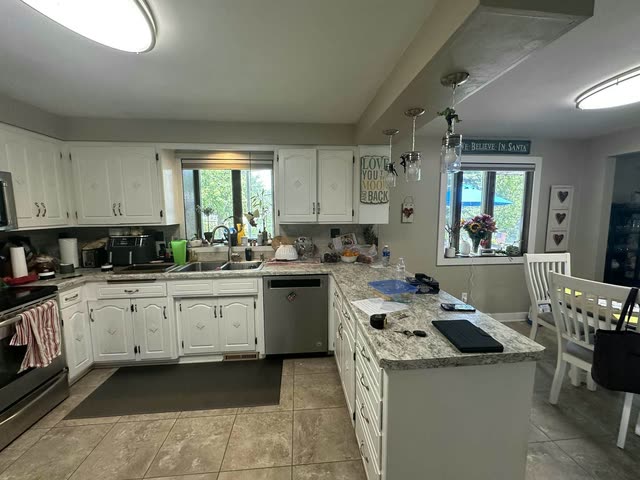 Before the remodel: Older kitchen with white cabinets with diamond accents, speckled laminate countertops, a peninsula under three jar pendant lights and a beige tile floor.