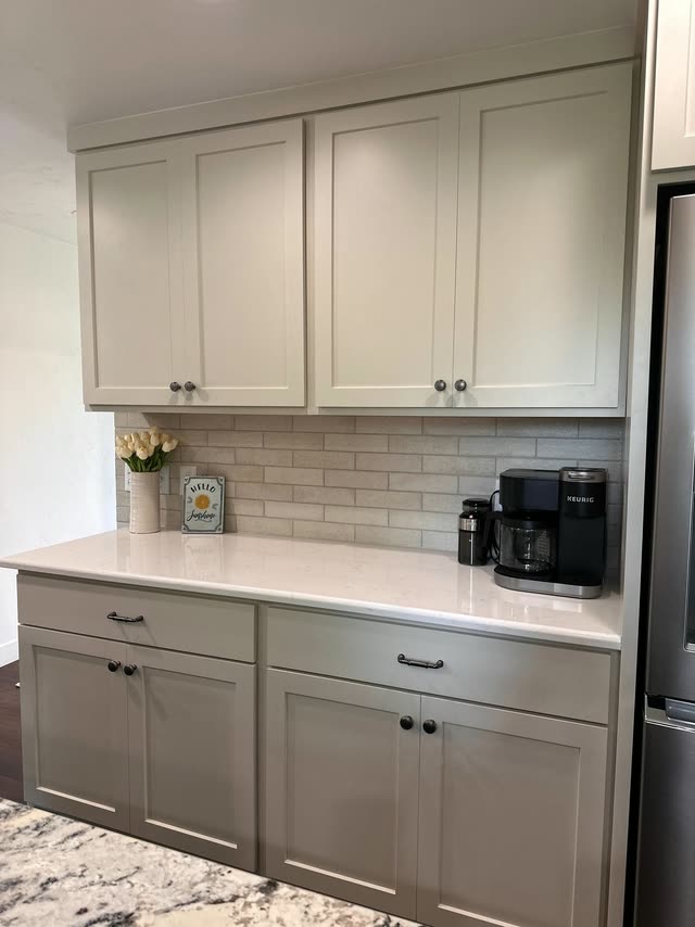 Straight-on view of greige shaker upper and lower cabinets with dark hardware, a white quartz countertop, textured tile backsplash, a vase of tulips and a coffee maker.