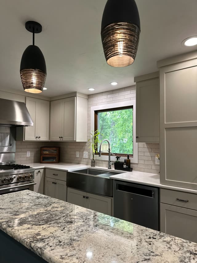 Remodeled kitchen seen across a granite island with a stainless apron-front sink, greige shaker cabinets, brick-look tile backsplash, a stainless range and hood and two black pendant lights.