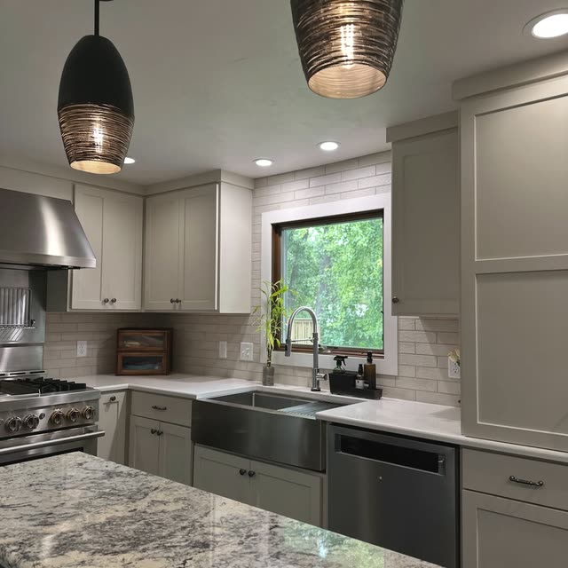 Kitchen with light greige shaker cabinets, a stainless apron-front sink under a window, a brick-look tile backsplash, a stainless range and hood, and two dark pendant lights over a granite island.