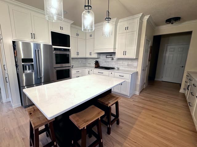 White shaker kitchen with a stainless French-door refrigerator, double wall ovens, a white range hood, a dark island with a white quartz top and three wooden saddle stools on light hardwood flooring.