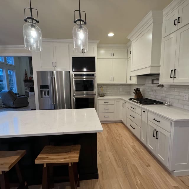White shaker kitchen with a quartz-topped island on a dark base, clear glass pendant lights, a stainless refrigerator and double wall ovens, a gas cooktop under a white range hood and light hardwood floors.
