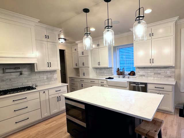 White shaker kitchen with a dark island topped in white veined quartz, three glass pendant lights, a gas cooktop with a pot filler, a farmhouse sink and light hardwood flooring.