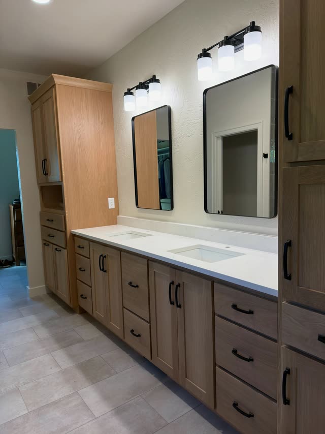 In progress: Double vanity in light natural-oak shaker cabinets with black pulls, a white quartz top with two undermount sinks, two black-framed mirrors and black vanity lights, flanked by tall linen cabinets.