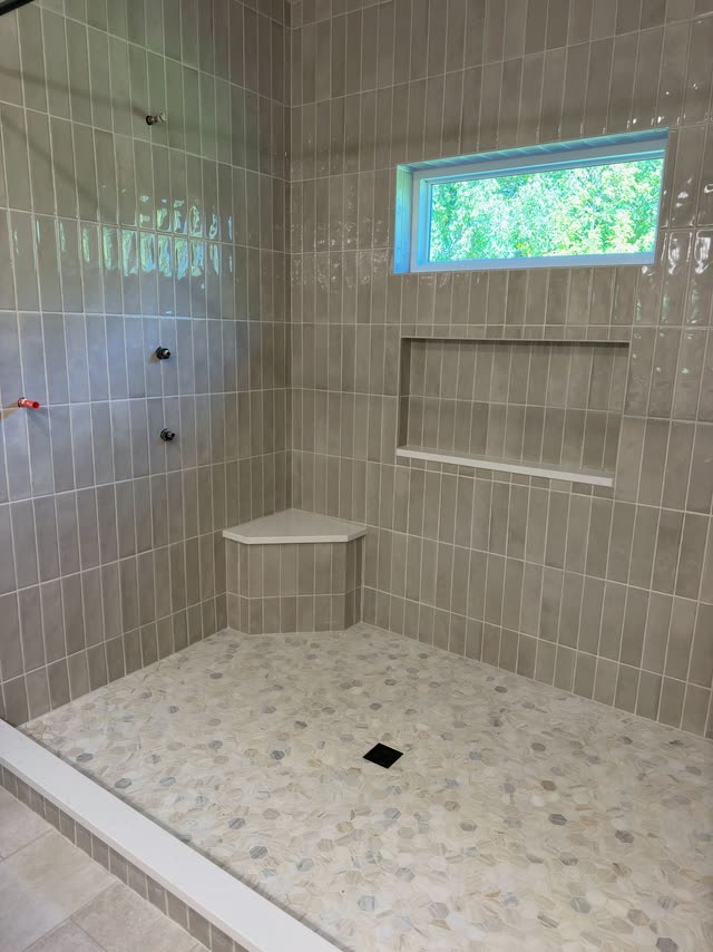 In progress: Large walk-in shower with glossy taupe vertical stacked tile, a recessed niche, a quartz-topped corner bench, a hexagon mosaic floor, a white quartz curb and a high window.