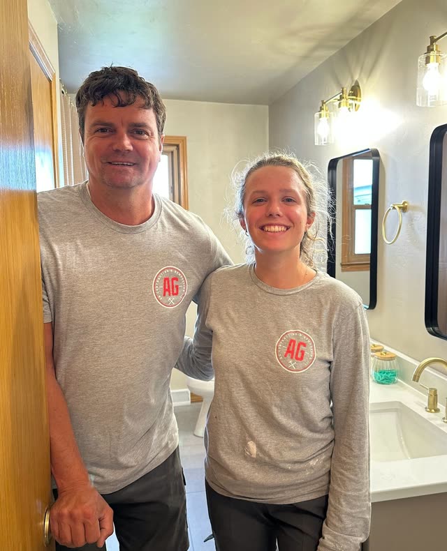 A man and a young woman in gray AG logo shirts smile in the doorway of a bathroom with a white vanity top, brushed gold faucet, black-framed mirrors and gold wall sconces.