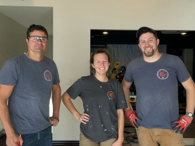 Three members of the AG Contracting team, two men and a woman in AG logo t-shirts, smiling on a job site with a miter saw behind them.