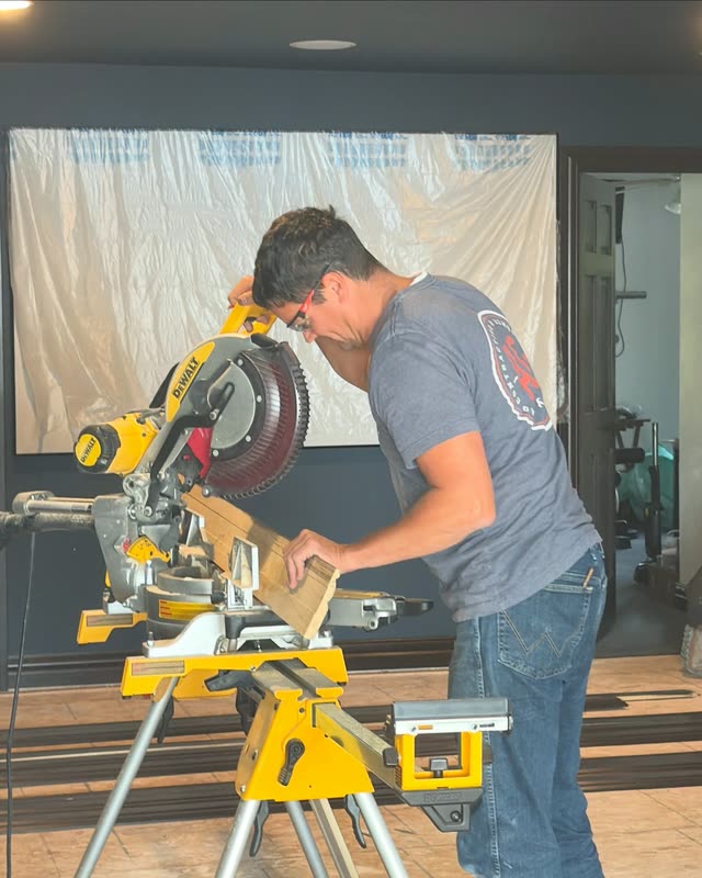An AG team member wearing safety glasses and a gray AG logo shirt cuts a board on a yellow miter saw in a room with navy walls.