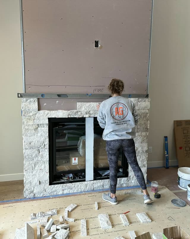 Team member in a gray AG Contracting & Remodeling sweatshirt, seen from behind, setting white stacked-stone veneer around a black fireplace insert.